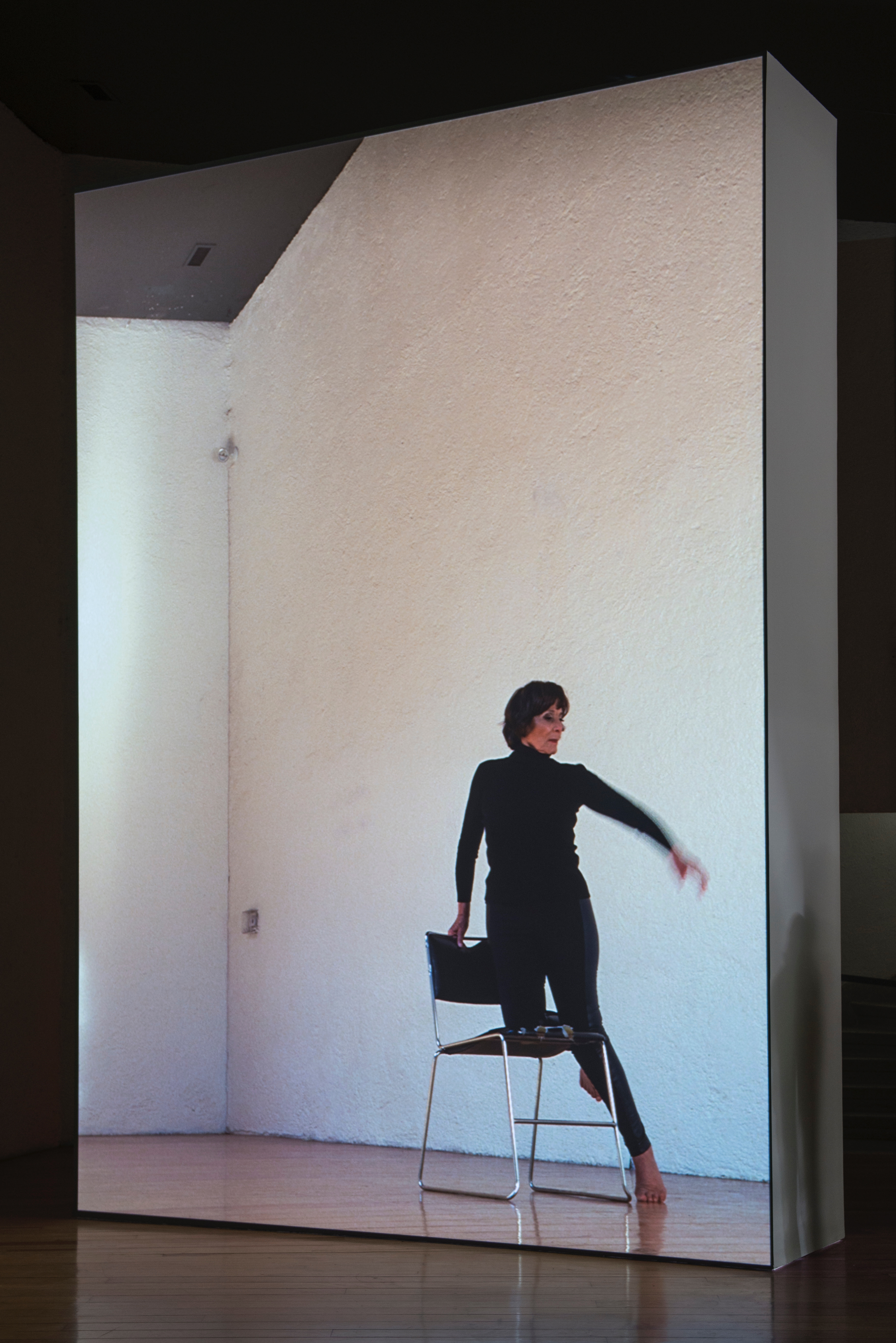 Installation photograph. A film is projected onto a floating wall. This still shows a woman wearing all black dancing, one knee bent onto a chair. Her other toes touch the wood floor, and one arm moves up, blurry in motion.