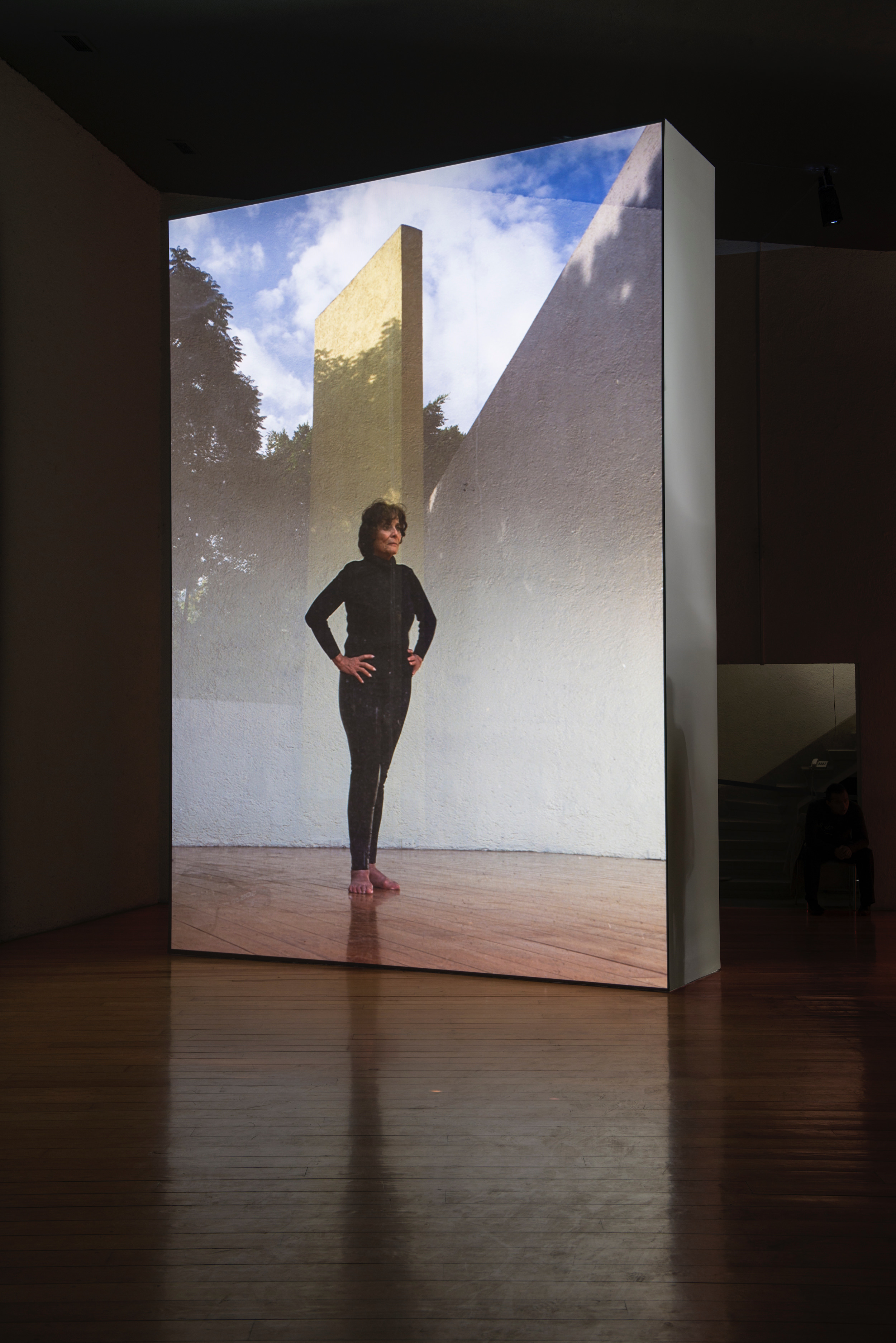 Installation photograph. A film is projected onto a floating wall. This still shows a woman wearing all black standing with her bare feet together and her hands on her hips. Projected behind her is a view of outdoor architecture or a sculpture.