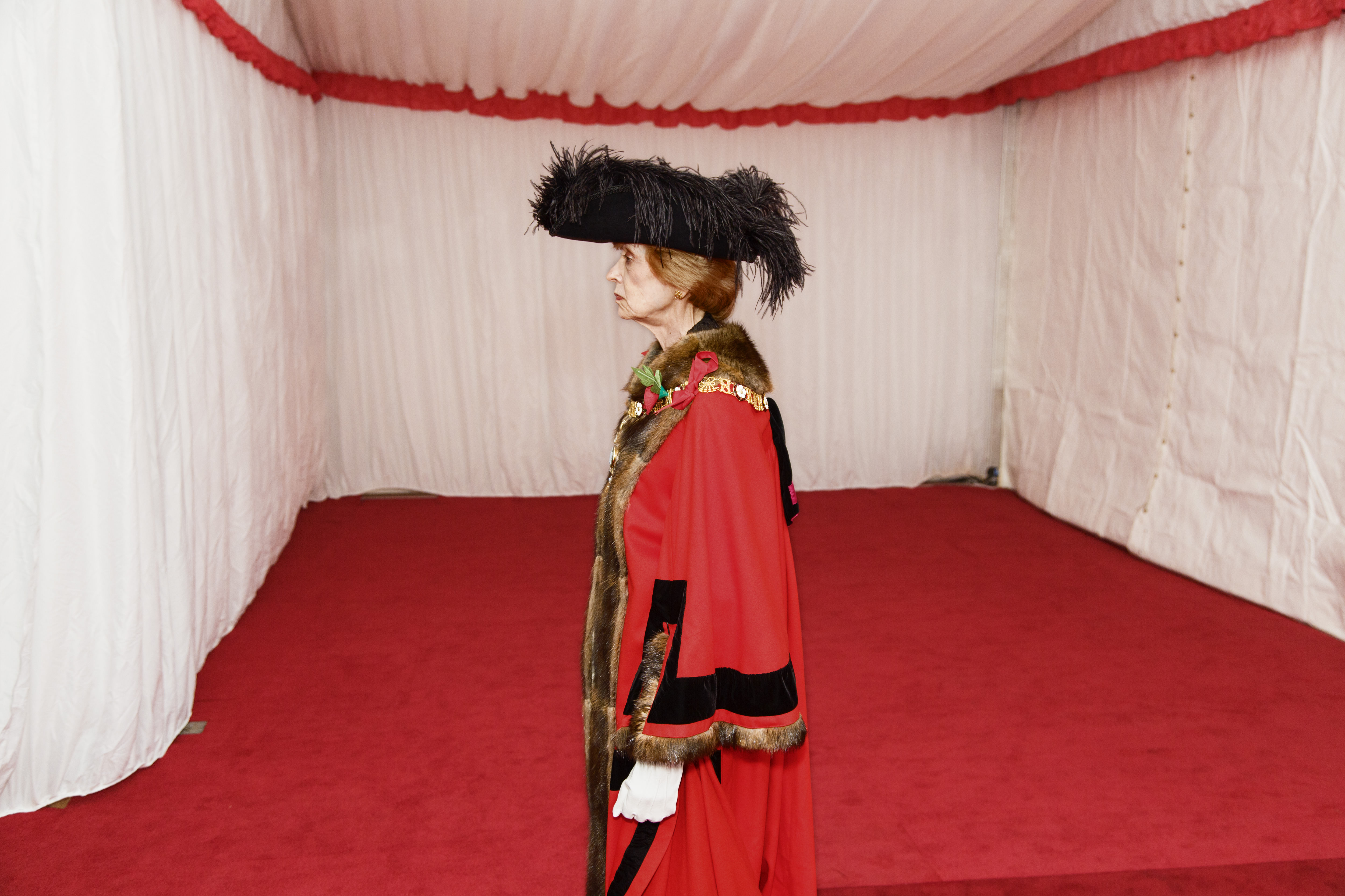 Colour photograph. Shown from the knees up, a woman wearing red regalia trimmed in black, gold, and fur faces our left in profile. Her black hat is lined with feathers, and the white-gloved hand we can see is in a loose fist at her side. She stands in what appears to be a white tent.