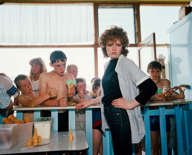 Colour photograph. A woman with hooded eyes under a poof of brown bangs looks at us, her downturned lips parted and her hand on her hip. She stands on our side of an ice cream counter, which is crowded with children on the other side.