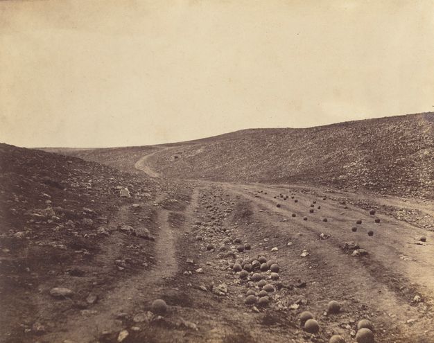 Black and white photograph. Dozens of cannonballs are strewn along a road and ditch in a shallow, barren valley.