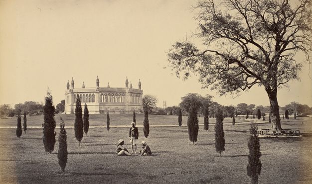 Sepia-toned photograph. Two men sit and one stands in a grassy park dotted with thin trees. The men have dark skin and wear light-coloured turbans. Across the field is an octagonal, temple-like structure.