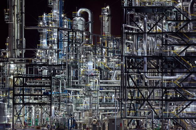 Colour photograph. Gleaming silver and jet-black pipes create an impenetrable web against a night sky. Lights among the equipment register as starbursts.