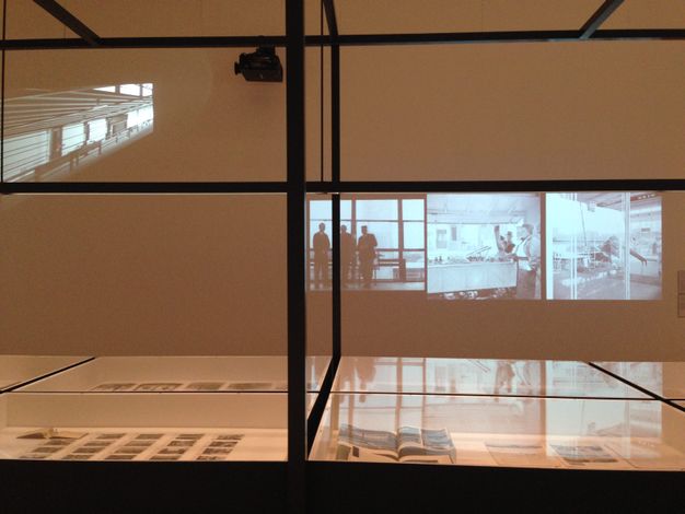 Installation photograph. Black and white films project over glass cases holding printed material and perhaps photographs.