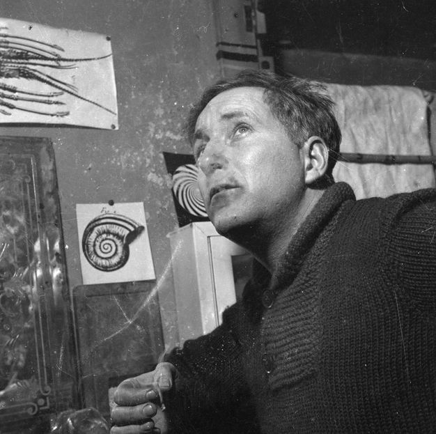 Black and white photograph. Shown from the chest up, a man’s body angles to our left as he looks sharply up under raised brows. Smoke wafts from a thin cigarette pinched between thumb and forefinger, along the bottom edge of the image. Works of art hang on the wall behind him.