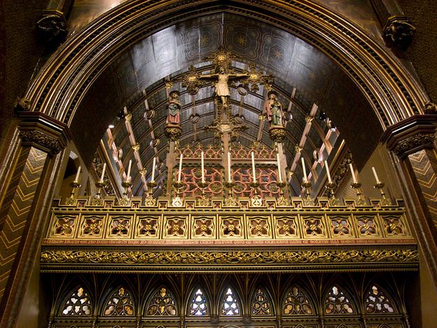 Rood screen. In this colour photograph, we look up at the top zone of a partition that fills an arch in a church. A row of candles lines several bands of gold tracery and ornamental patterns. A crucifix rises into the arch above, and is flanked by two people, the Virgin Mary and St John the Evangelist, standing on miniature platforms to either side.