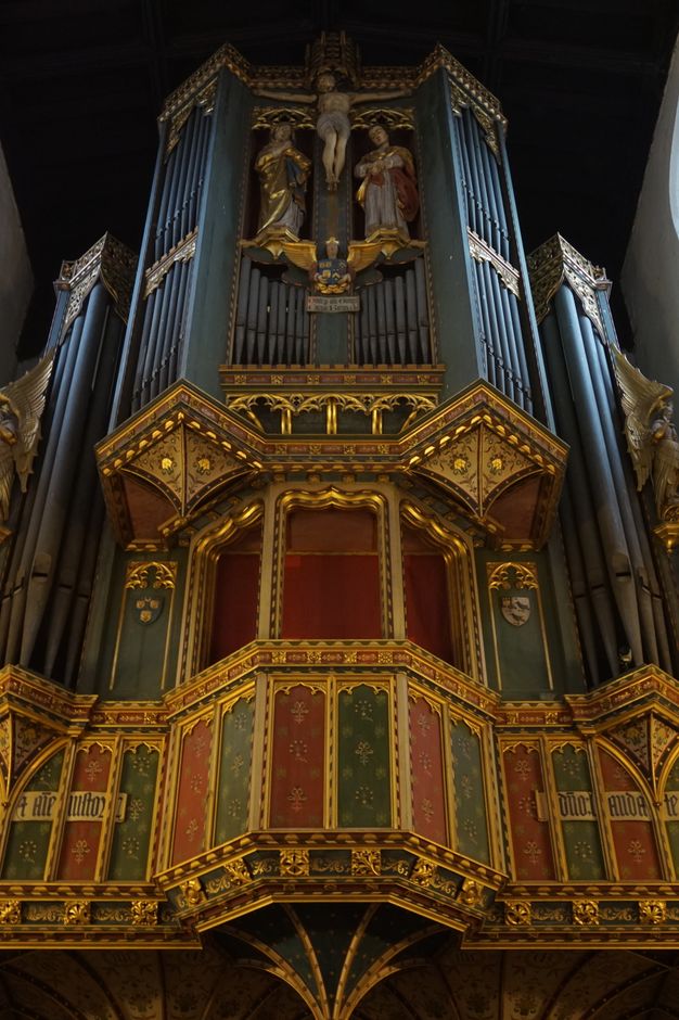 Colour photograph. The pipes of a church organ soar into the shadowy vault above. It rises from red and green panelwork, with painted gold ornamentation. A crucifixion with the Virgin Mary and St John the Evangelist to each side is tucked among the pipes at the top centre.