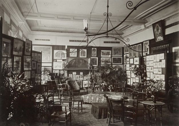 Black and white photograph. The walls of a room are hung densely with paintings and works of art. About a dozen chairs, sofas, and small tables are set up in the space. Sprays of potted plants add to the cluttered feeling.
