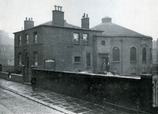 Black and white photograph. A blocky building with at least three chimneys is attached to a round structure like an oversized turret. A wall separating us from the structure is half as high again as a person walking on the pavement next to it.