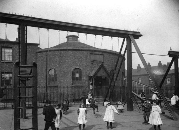 Black and white photograph. Girls in white frocks and boys in dark school uniforms swing on tall swings or play in a paved courtyard outside a round brick building with a peaked roof.