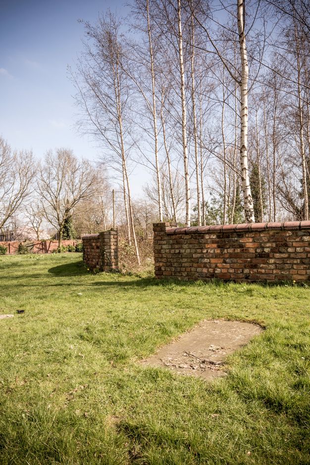 Colour photograph. An opening in a rounded brick wall leads to a copse of spindly trees. A rectangular patch of dirt mars the grassy field that surrounds the wall.