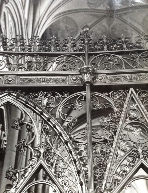 Choir screen. This black and white photograph focuses on the intricately swirling ironwork along the top edge of a choir screen. The vaulted ceiling of the church is glimpsed above.