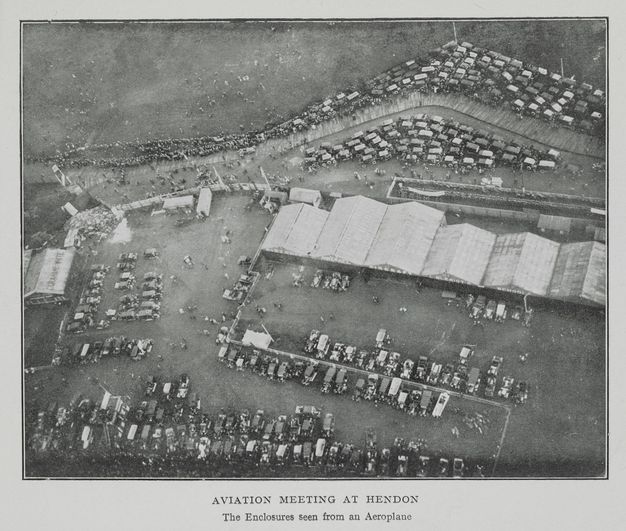 Black and white photograph. A high, aerial view of flat grounds. Six wide hangars are lined up near the centre of the composition. Cars are parked around the edges of the fields, and people dot the landscape. Text below reads, “Aviation Meeting at Hendon The Enclosures seen from an Aeroplane”.