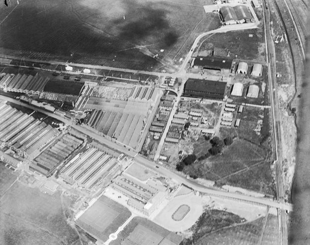 Black and white photograph. A high, aerial view onto a complex of buildings like warehouses or hangars, which together make a roughly backwards-facing check mark. The image is slightly blurry, and clouds cast shadows on the ground in the upper left corner.