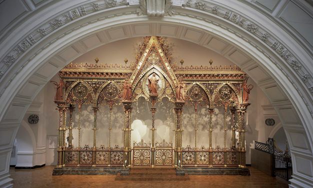 Choir screen. In this colour photograph, we see a freestanding architectural partition, like a floating wall, has five bays, each under a pointed arch. Each arch is then bisected by a spindly column. The central bay is the widest and tallest, and a person stands at its apex. Intricate tracery encrusts the upper edges of the screen and fills in the arches. Panels across the bottom are also made with intricately patterned tracery. Most surfaces are shiny or burnished gold, and there are also touches of muted red, warm yellow, and earthy green.