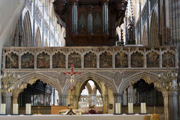 Architecture. Colour photograph of a choir screen in a large church. Three arches support a row of eleven paintings in a gallery-like level. Organ pipes above the screen are backlit by light pouring into the transept beyond. The arches are wide and slightly pointed, and the spandrels in between are intricately carved with leafy designs.