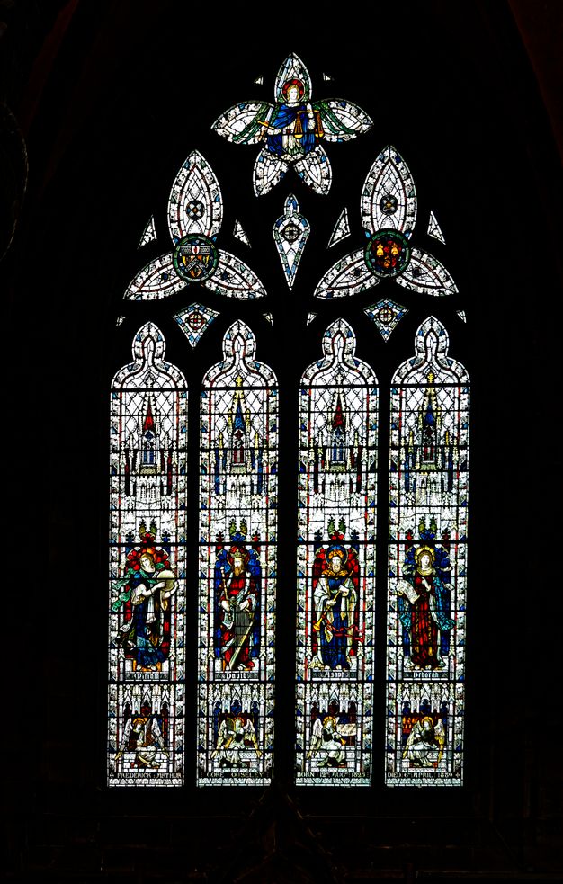 Stained glass window. Four people stand, each in a tall narrow pane, and each holds a musical instrument. The four panels are topped with trefoil and five-lobed windows. Flecks and slivers of glass between the larger components glint like sparks against the black surround.