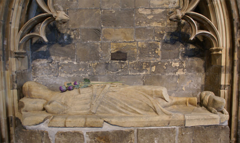 Sculpture. A carved effigy shows a man wearing robes lying on his back on a slab set in an arched niche decorated with tracery. One leg is broken off below the thigh.