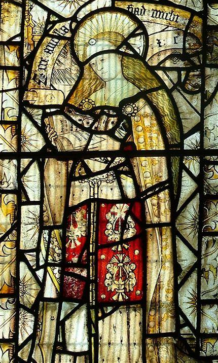 Stained glass window. Detail view of a haloed woman with long blond hair, the Virgin Mary, reading a book. Pieces of glass around her have sunbursts overlaid with blooming white roses.