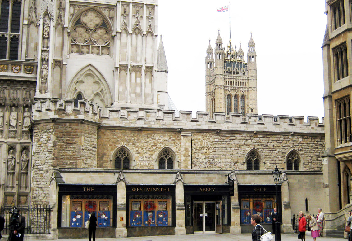 Architecture. A two-storeyed, hall-like stone structure stretches across a church façade. The second level has two-part, pointed arched windows and crenelation along the roofline. The lowest level has storefront windows and the sign, “The Westminster Abbey Shop”.