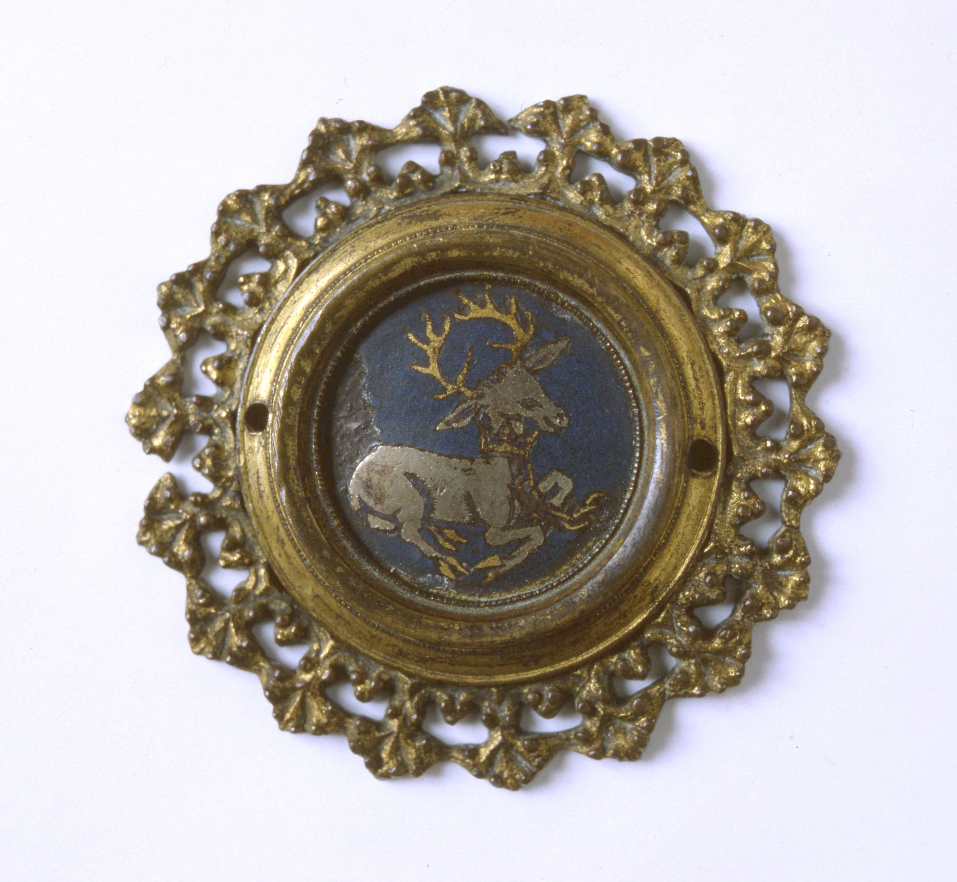 Plaque. At the centre of this medallion-like roundel is a silver stag with gold antlers lying on a blue field. The stag has a gold crown around its neck with a gold chain attached to it. Flaring, gold-coloured leaves create a ruffled edge around the perimeter.