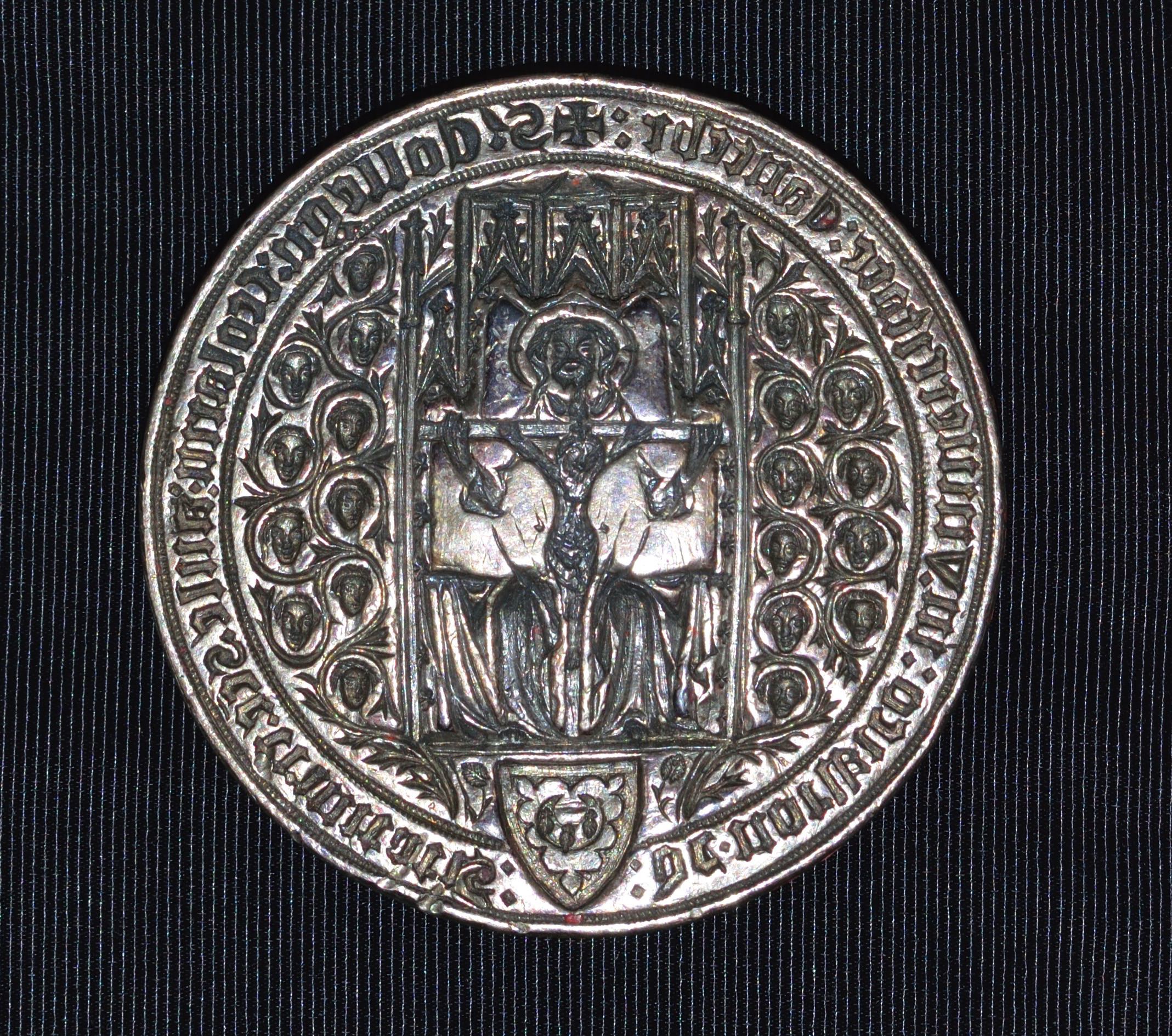 Seal. In this round seal, an enthroned man, God the Father, holds a crucifixion. Twenty miniature heads, ten to each side, are encircled with continuous vines sprouting near a coat of arms. Gothic writing runs around the perimeter, in reverse.