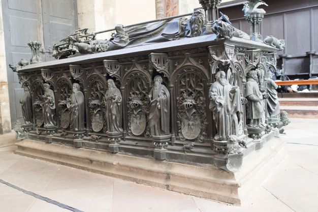 Sculpture. The sides of this rectangular bronze tomb are decorated with arches filled with tracery and heraldic crests. Between each arch and the next is a figure standing on a hexagonal platform under a miniature canopy. Those on the sides facing us appear to be all men wearing robes, and they hold one or two objects. They have curly or wavy hair, and most have full beards. We are low to the ground so only catch a glimpse of the figure on the top of the tomb.