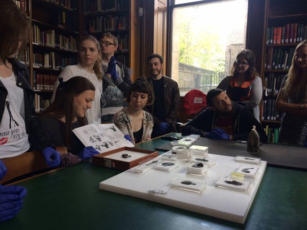 Colour photograph: Nine men and women gather around a table laid with artefacts in a book-lined room. Many look at a book held open by a woman sitting at the table. She and others wear blue gloves.