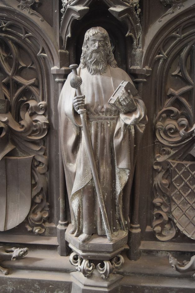 Sculpture. A man with a long beard and hair, and wearing robes belted at the waist, stands holding a closed book in one hand. In the other, he holds a staff with a knob at the top and a large shell below it. The hexagonal platform on which he stands is supported on leafy flourishes. The surfaces are muted, silvery gold.