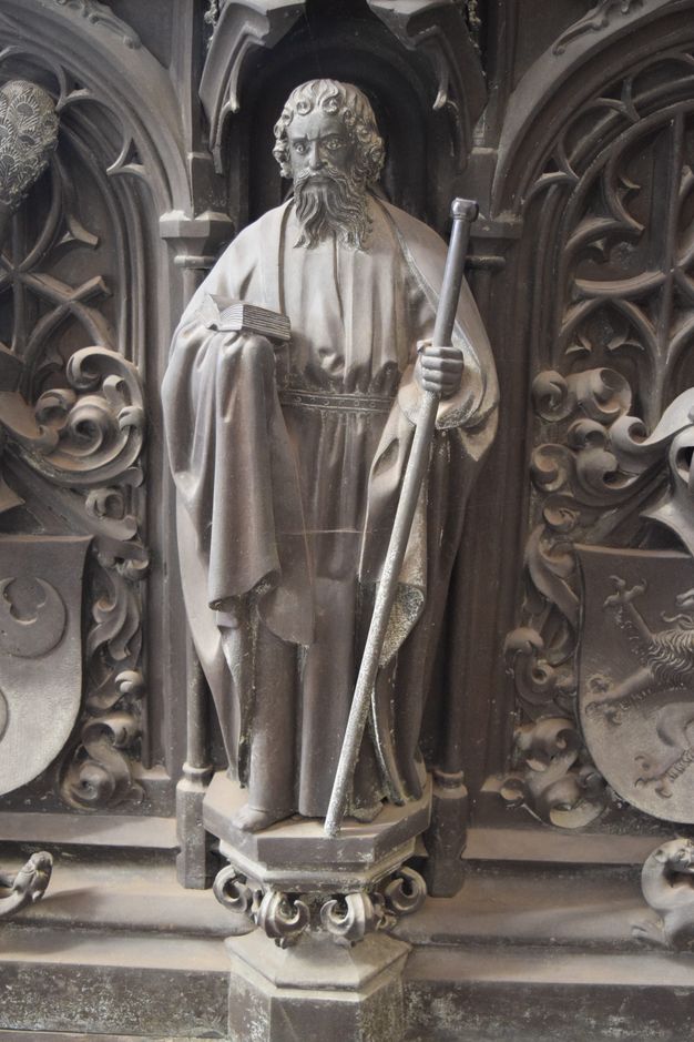 Sculpture. A man with curly hair and a long, forked beard wears robes belted at the waist. He holds an open book in one hand and a staff with a knob at the top in the other. The hexagonal platform on which he stands is supported on leafy flourishes. The surfaces are muted, silvery gold.