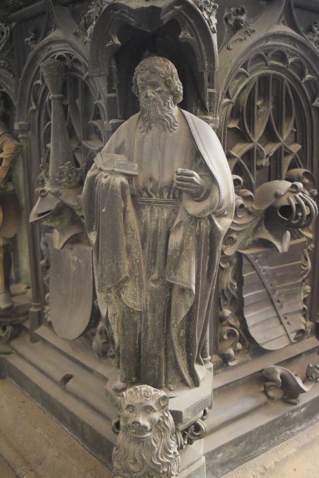 Sculpture. A man with curly hair and a long, forked beard wears robes belted at the waist. He holds an open book in one hand. His other hand is empty and held before him as if grasping something that is no longer there. The hexagonal platform on which he stands has a lion at the front. The surfaces are muted, silvery gold.