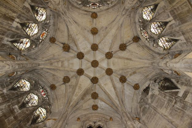 Architecture. We look up at a ribbed grey stone ceiling which has a hexagonal shape and stained glass windows on several sides. Knots of decorative foliage sit at each site where the ribs intersect.  