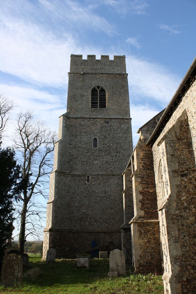 Architecture. A four-storey, wide stone tower rises over a porch projecting from the side of a church. In this view, the right edge, facing the church, is straight, and the front left corner has stepped buttresses. Small windows pierce the second and third levels, and a larger opening is on the top storey, which is crenelated.