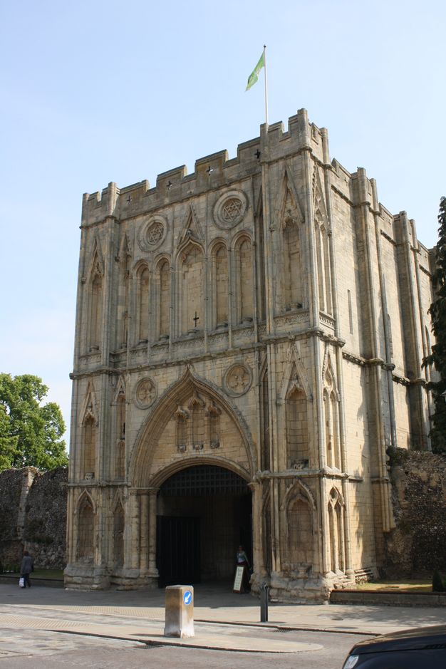 Architecture. A squat stone structure like an elongated square rises above a wide, shallowly arched opening on the ground level. Blind arches are spaced across the two tall storeys above. A person walking out the door shows that the arched opening alone is at least two typical storeys high.