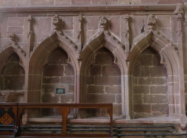 Architecture. Four niches under pointed arches are backed by pale red stone. The arch over each niche comes to a point and is topped with a finial. The niche to our left is smaller, perhaps two thirds, the size of the others.
