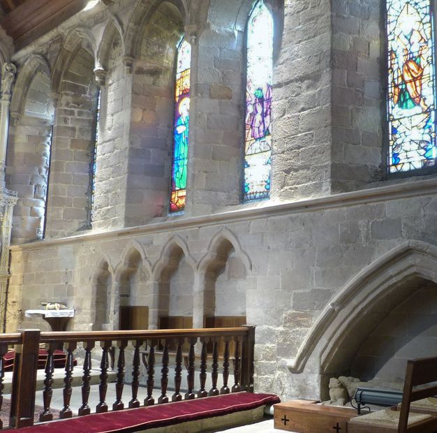 Architecture. A row of four, three-lobed blind arches, each with a wooden seat, are below a level of arched stained glass windows set into the grey stone walls of this church.