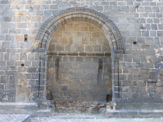 Architecture. A round topped, arched, shallow niche is set into a grey stone wall. The bench, which would have acted as the seat, has been damaged or removed.