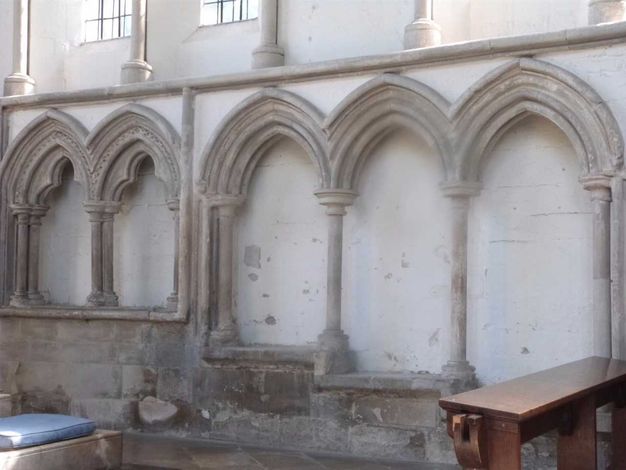 Architecture. Three pointed arches outlined in grey stone against a white wall create three niche-like spaces. A wide ledge below steps down in each successive niche, resulting in seats at three levels.
