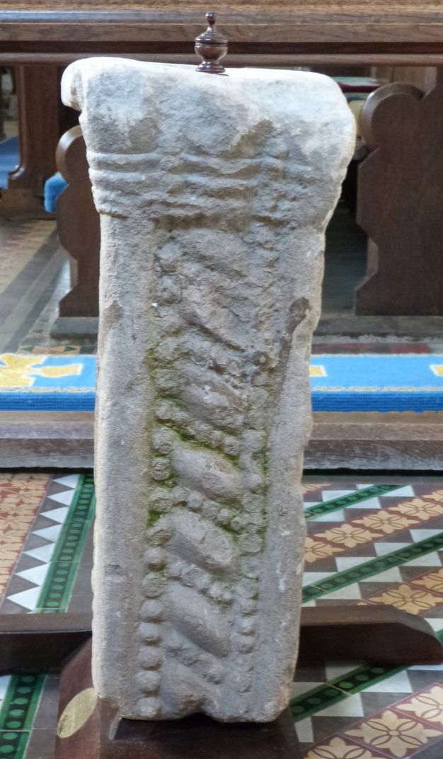 Architectural feature. A fragment of a grey stone, square pillar is carved with a geometric design and is topped with a wooden finial.