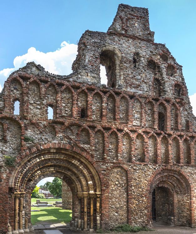 Architecture. A partially ruined exterior wall made of irregular stonework and red brick detailing has two doorways with archivolts below two levels of blind arches.