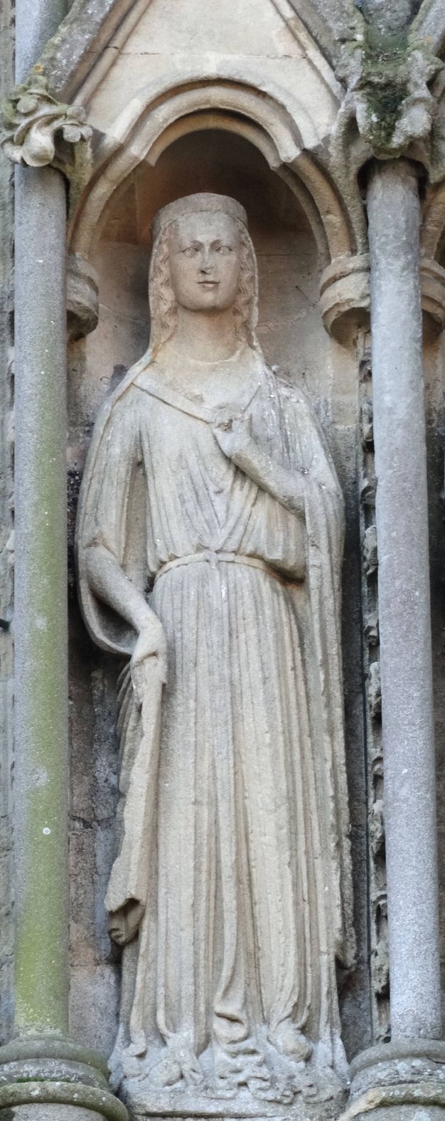 Sculpture. A statue of a woman wearing a veil and a long dress stands in a niche on the exterior of a church. She holds one hand up to her chest and holds the veil or a cloak with her other hand.