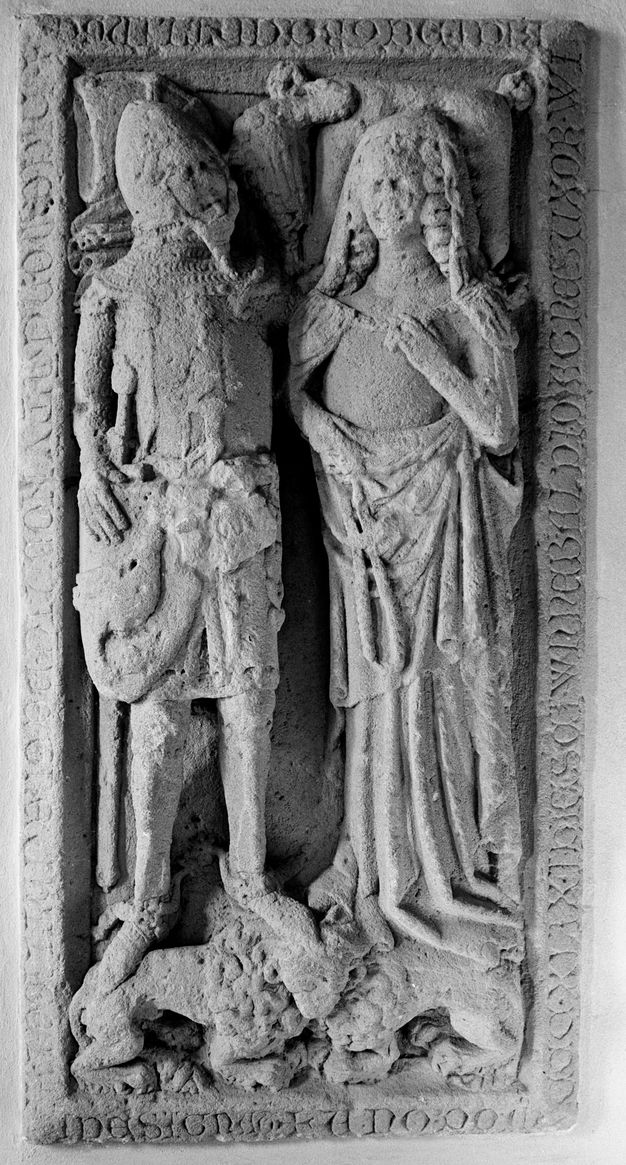 Sculpture. We look down on a carved monument showing a man and woman lying on their backs alongside each other. The man wears armour and the woman a veil and long dress. Their feet rest on the backs of lions at the foot of the bier. The man’s head turns towards the woman and his arm extends under her shoulders.
