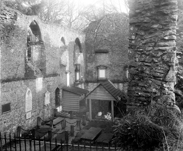 Black and white photograph. Inside the partially ruined choir of a stone church, a wooden structure near the far wall covers a carved monument showing two people lying alongside each other.