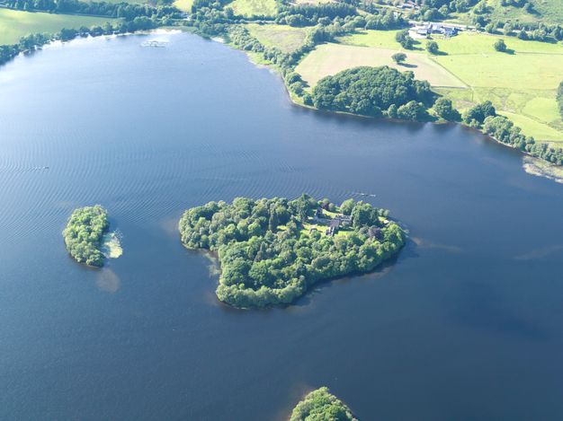 Colour photograph. A lightly rippling blue lake surrounds a small island with at least one building. An even smaller island is nearby, and the corner of a third peeks in from the lower edge. Trees line the shoreline along the far side of the lake.