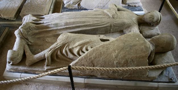 Sculpture. A carved monument shows a man, Walter Stewart, closer to us, lying alongside a woman, Mary Stewart, who turns her face to look at him. Mary’s long dress covers her legs and feet. One arm reaches towards Walter. He lies on his side, facing her. The surface is rough, and some details, especially in their faces, are damaged or lost.