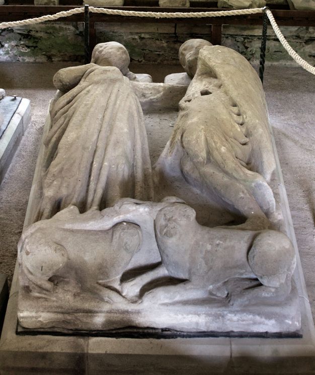 Sculpture. From near their feet, we look at the carved monument of a man and woman lying next to each other. Two dogs are underfoot. The woman lies on her back and the man on his side.