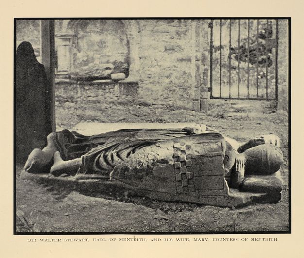 Black and white photograph. A monument showing a man and woman lying next to each other lies on the ground outside. Closer to us, the man lies on his side with a shield at his back. The woman’s hand reaches for him and cups the back of his neck.