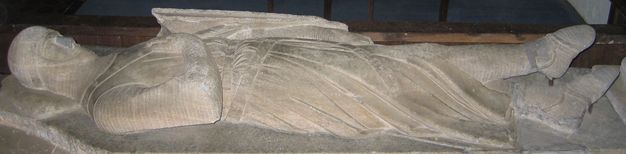 Sculpture. Carved from stone, a knight wearing armour lies on a slab, his shield on one arm. The other hand rests on his chest.