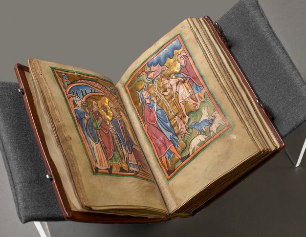 Pages of a book. A leather-bound volume rests partially open to reveal two illuminated parchment pages. The scene to the left shows a man and a woman in a temple facing a priest. The scene to the right shows an angel giving a message to a man in a cloak and pointed hat. Both scenes are painted with saturated, jewel tones in blue, pink, green, and white, and bright gold.