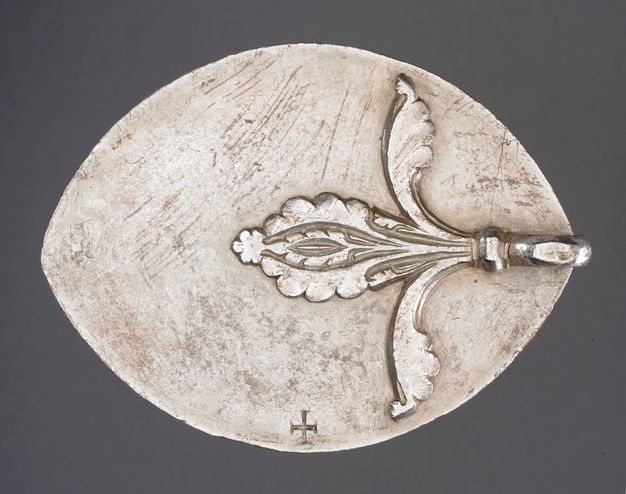 Reverse of a seal. A silver oval with pointed ends has a small handle topped by a fleur-de-lis-shape in relief attached at the right-hand point. A cross is engraved at the bottom centre of the seal.